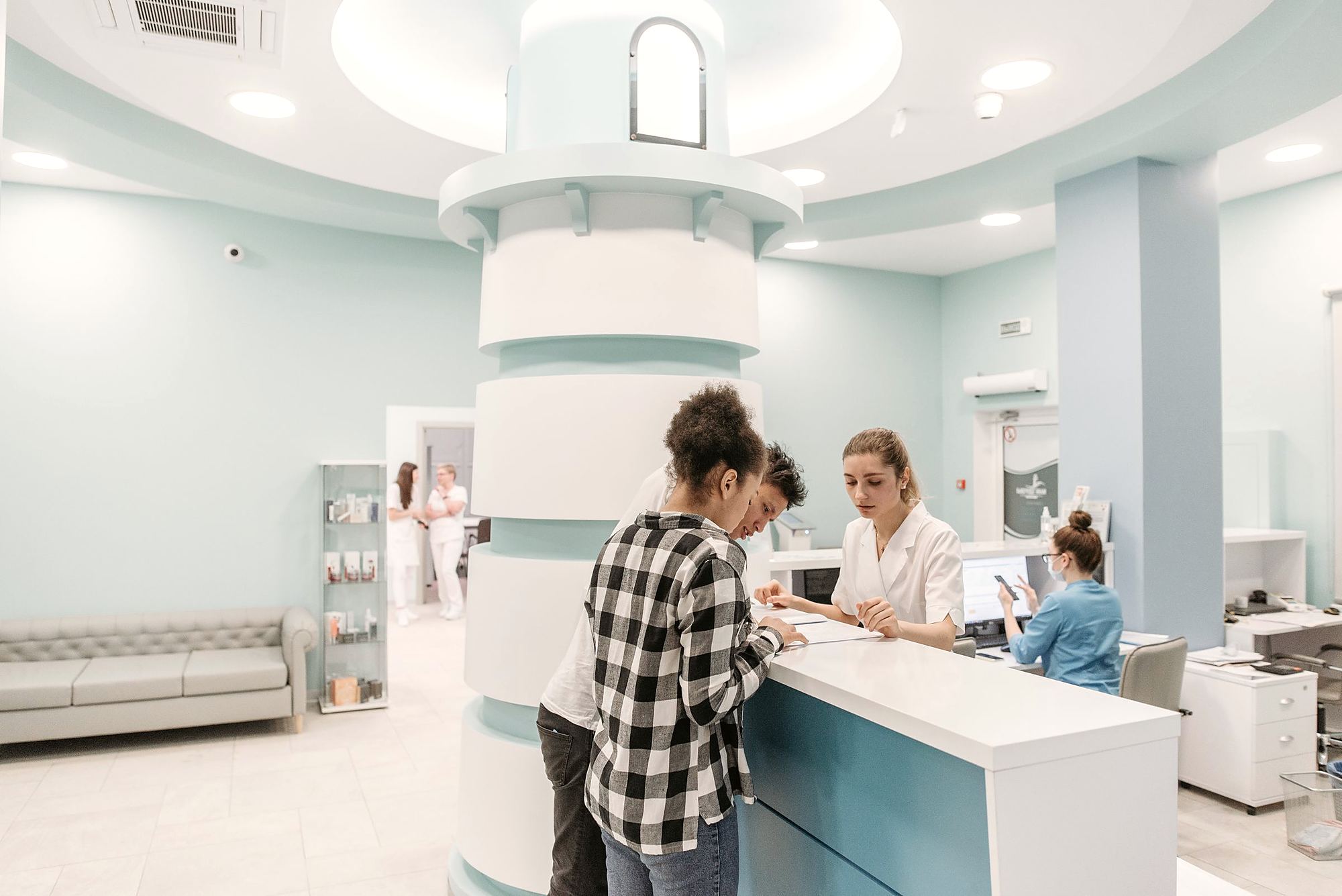 A busy clinic reception: a receptionist helping two patients register while staff work in the background